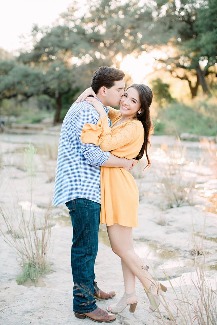 Bride smiles as her fiance kisses her on the cheek for their engagement photos at Bull Creek