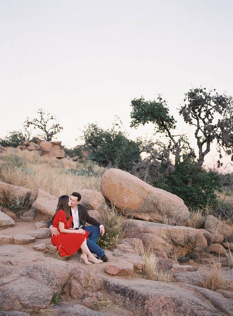 Girl in a red dress and man in a blue suit sit at enchanted rock for engagement photos