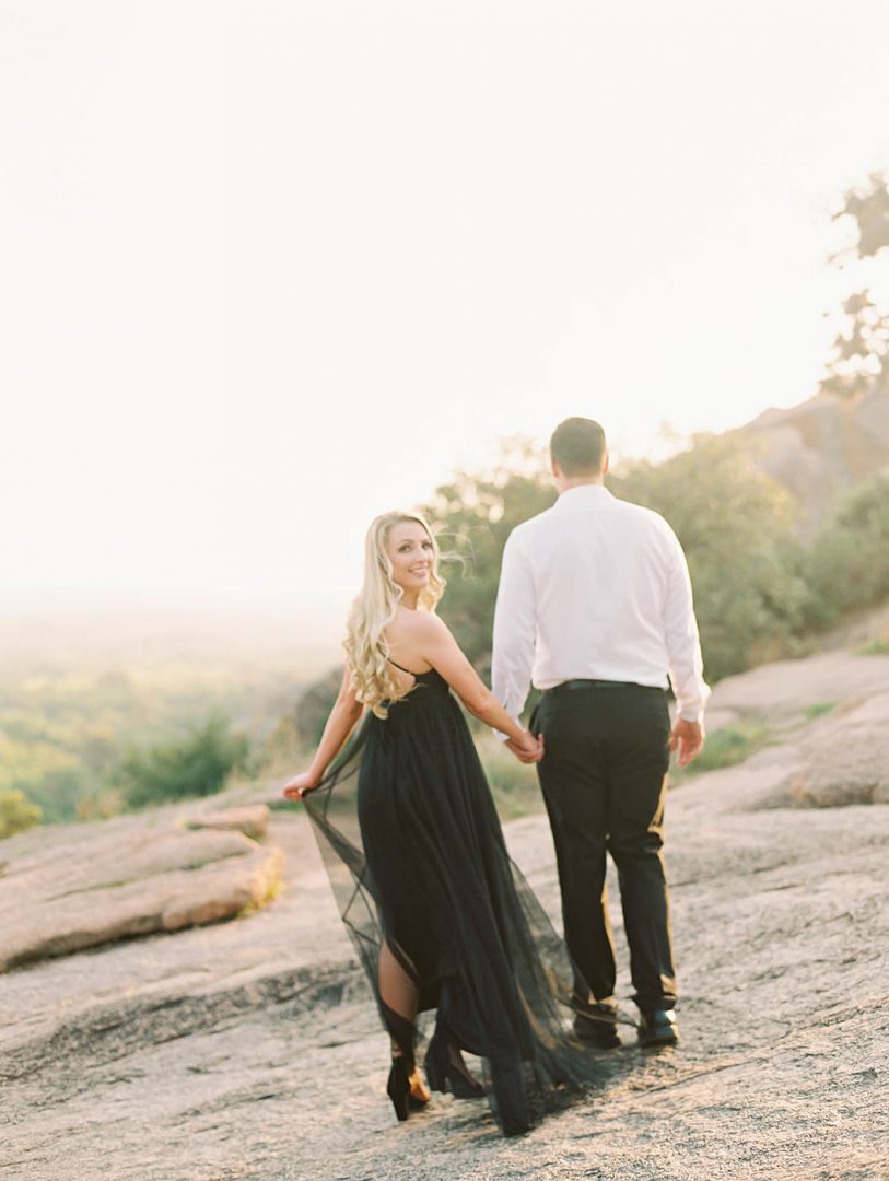 Engagement photo of a girl in a black dress looking over her shoulder while walking with her fiance