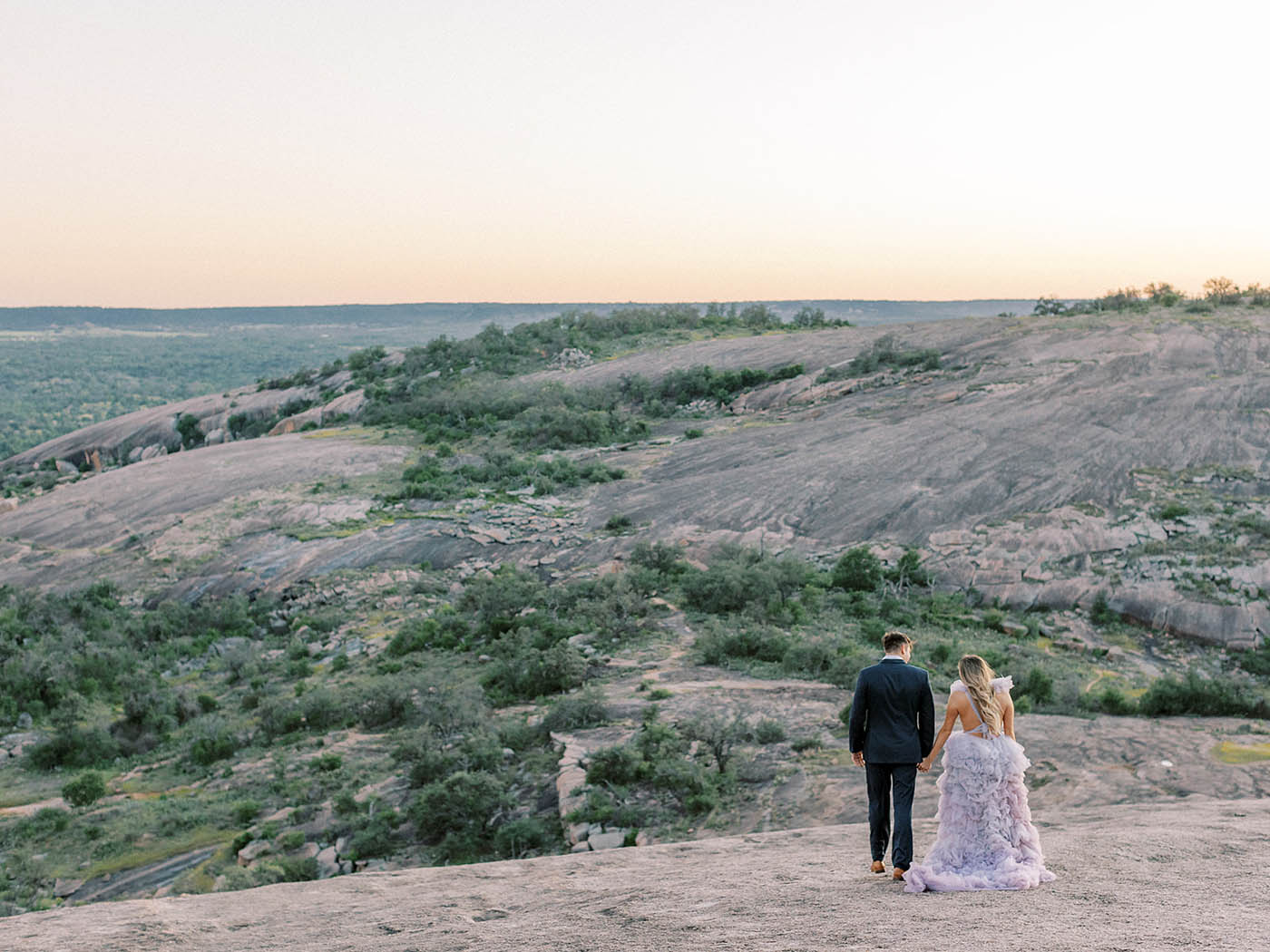 wide shot of a couple in a suit and ruffled dress walking down enchanted rock after sunset
