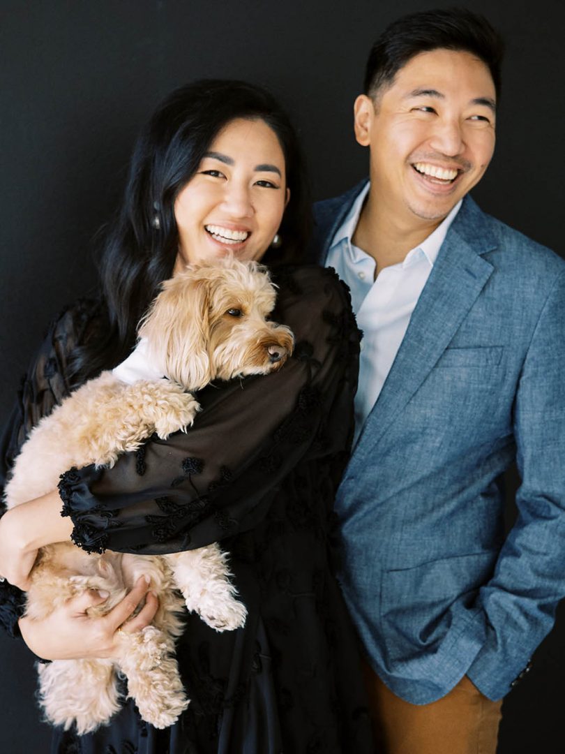 Couple poses against a black wall with their dog for engagement photos