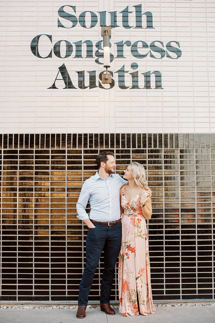 Couple poses together against a white tile wall outside the South Congress Hotel