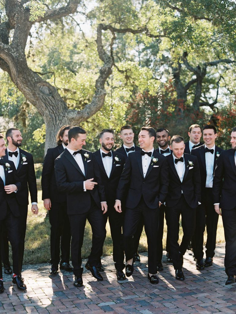 The groom and groomsmen laugh together in black tuxedos