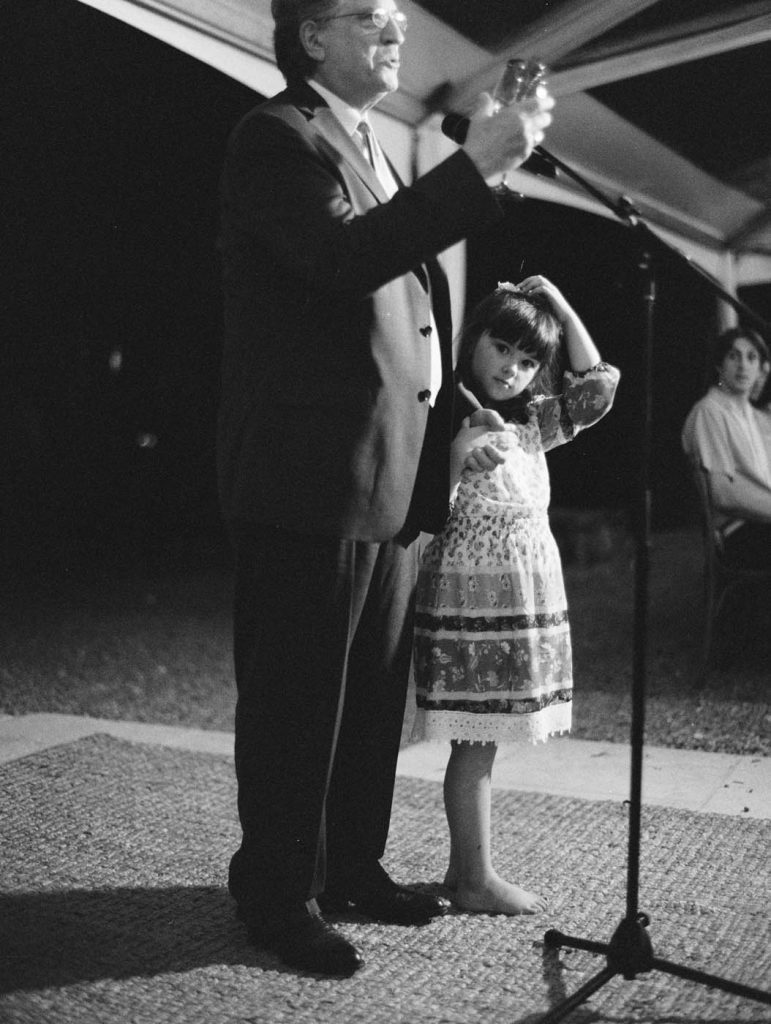 a child stands next to her grandfather as he gives a toast at the wedding