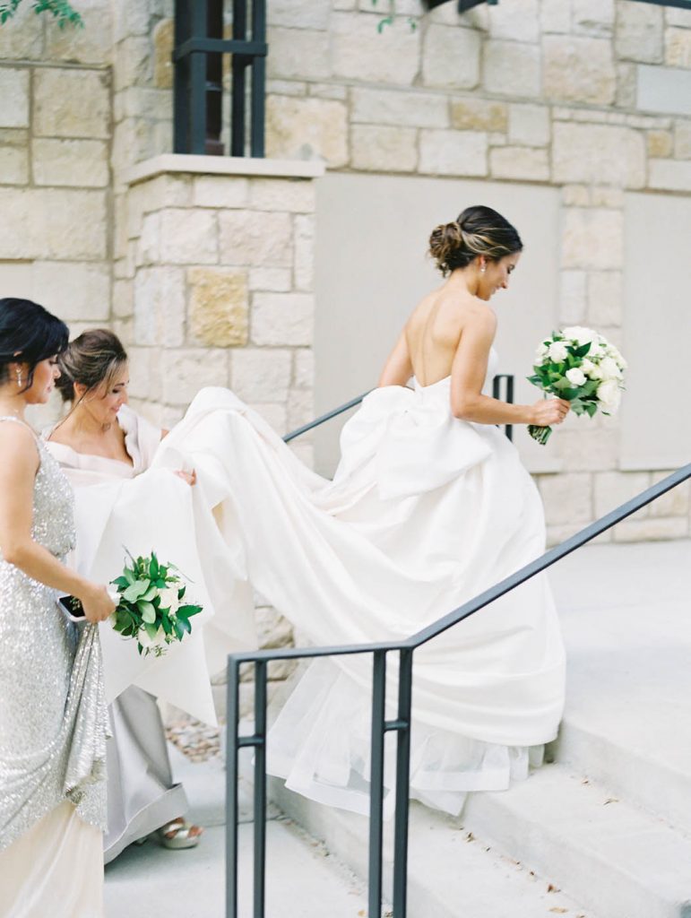 bride walks up the stairs with mom and bridesmaids carrying her train
