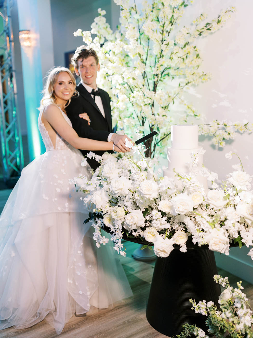 the bride and groom cut their modern white cake surrounded by colorful lighting