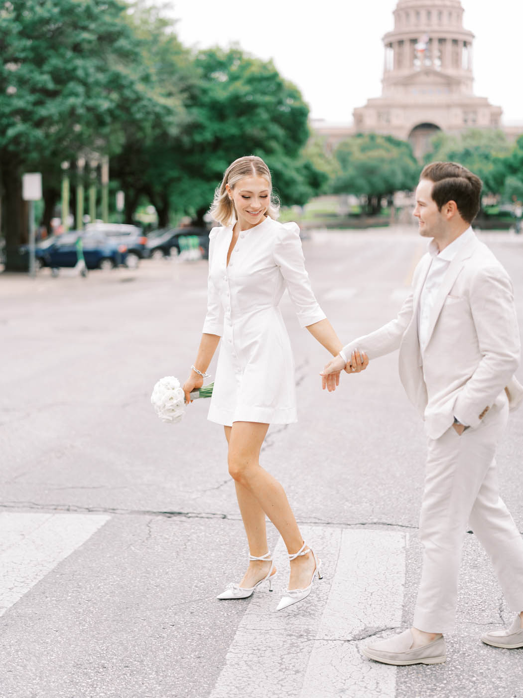 woman in a short white dress and Mach and Mach heels leads her new husband across an intersection with the Austin capital in the background