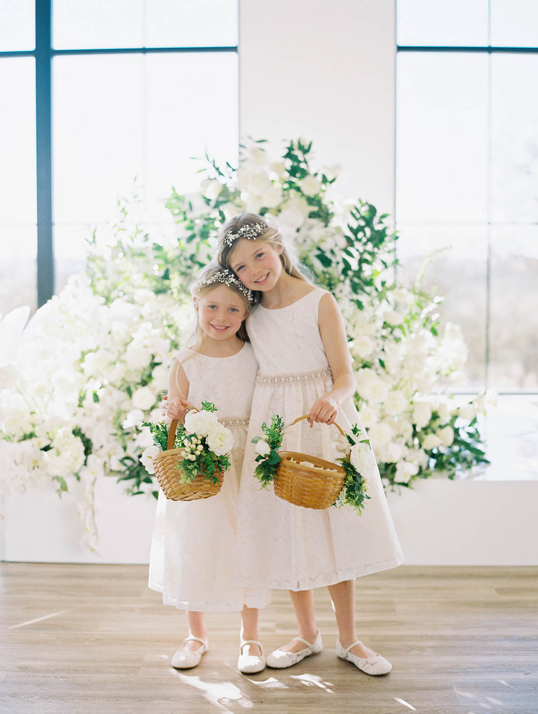 the flower girls smile together and hold their baskets in front of the piano decked out in white and green floral